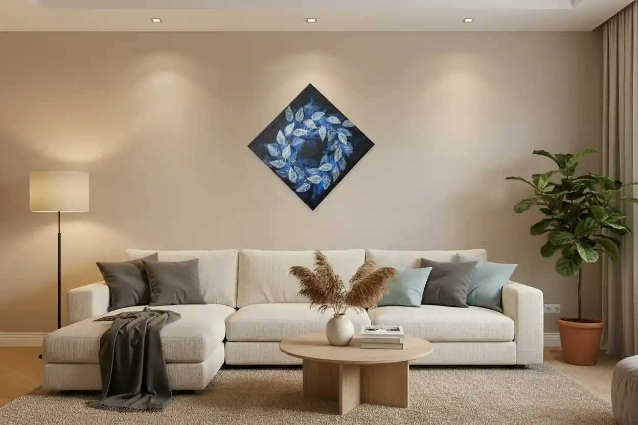 A modern living room featuring a diamond-shaped canvas wall art print with a blue leaf wreath design, hung above a white sectional sofa with neutral-toned pillows and a round wooden coffee table.