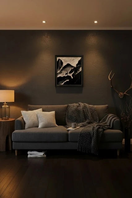 A dark gray fabric sofa in a moody living room setting, styled with two textured white throw pillows, a chunky knit gray throw blanket, and a black-framed black and white mountain landscape print hanging on the dark gray wall above it.