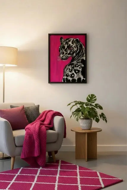 A framed art print of a spotted big cat on a vibrant magenta background, styled in a modern living room setting above a gray armchair with a pink throw blanket and a small potted plant on a wooden side table.