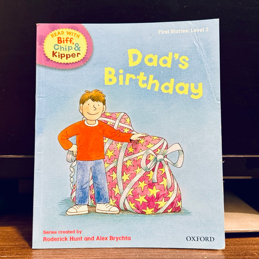Children's book titled 'Dad's Birthday' by Biff, Chip, and Kipper. A boy is next to large gift, on a blue background.