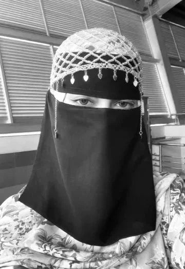A black niqab face veil worn by a woman, paired with a white and black beaded skullcap, shown in a black and white close-up portrait.