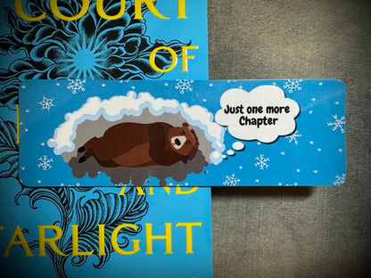 A winter themed laminated card stock bookmark with a picture of a hibernating bear on a blue background with a quote saying 'just one more chapter'. placed on top of a light blue colored book.