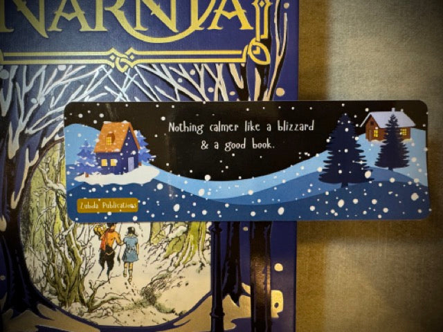 A colorful winter-themed bookmark with the quote 'nothing calmer like a blizzard & a good book' spread out on a surface with Narnia in the background.
