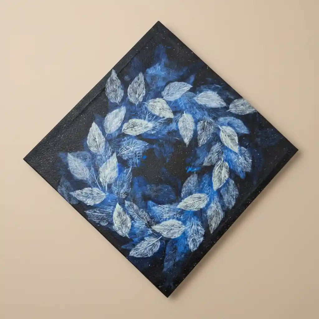 A square canvas painting shown at an angle, featuring a circular wreath of textured blue and white leaves on a dark navy and black background, mounted on a plain beige wall.