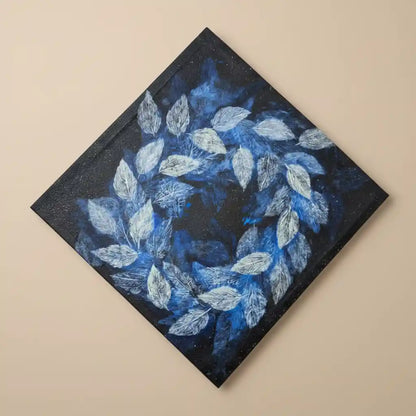 A square canvas painting shown at an angle, featuring a circular wreath of textured blue and white leaves on a dark navy and black background, mounted on a plain beige wall.