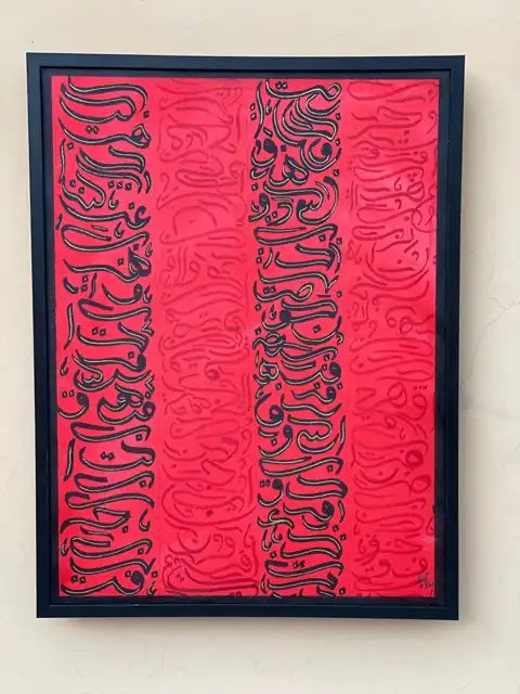 Framed wall art featuring a vivid pink-red background with three vertical columns of black Arabic calligraphy, mounted on a light beige wall.