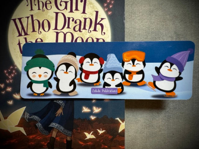 A colorful winter-themed bookmark with skating penguins spread out on a surface with the book the girl who drank the moon in the background.