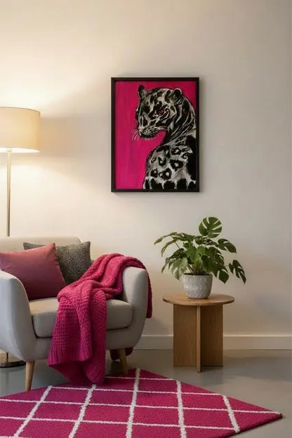 A framed art print of a spotted big cat on a vibrant magenta background, styled in a modern living room setting above a gray armchair with a pink throw blanket and a small potted plant on a wooden side table.