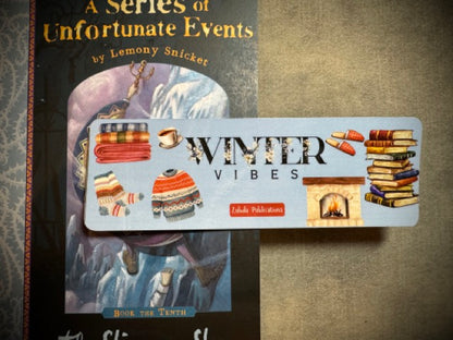 A colorful winter-themed bookmark with the words 'Winter vibes' spread out on a surface with the book a series of unfortunate events in the background.