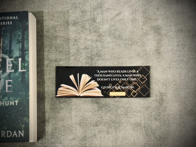 A  laminated card-stock bookmark with an open book and quote that says 'A man who reads lives a thousand lives; A man who doesn't lives only one' on a black background placed on a surface with the wheel of time book in the background.