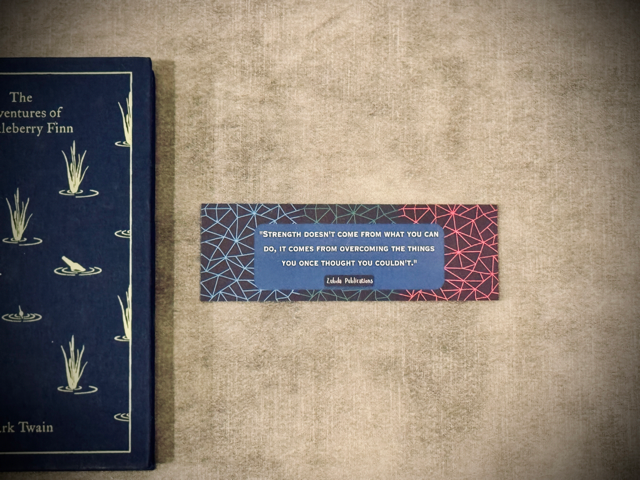 a laminated card stock bookmark with the quote 'strength doesn't come from what you can do, it comes from overcoming the things you once thought you couldn't' on a patterned background. Placed next to The Adventures of Huckleberry finn book