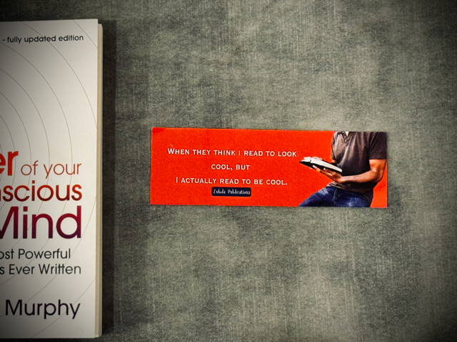 A  laminated card-stock bookmark with a man holding a book and bright orange background placed on a surface with the book 'power of your subconscious mind' in the background.