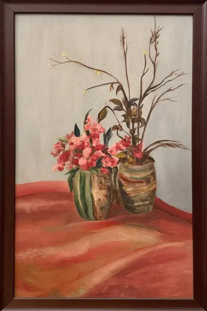 A framed still life painting featuring pink flowers in two textured vases on a draped red-orange surface against a light gray background, enclosed in a dark brown wooden frame.