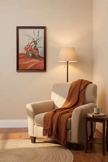 A framed floral still life painting hangs on a beige wall above a cream armchair with a rust-orange throw blanket and a floor lamp in a warmly lit living room corner.