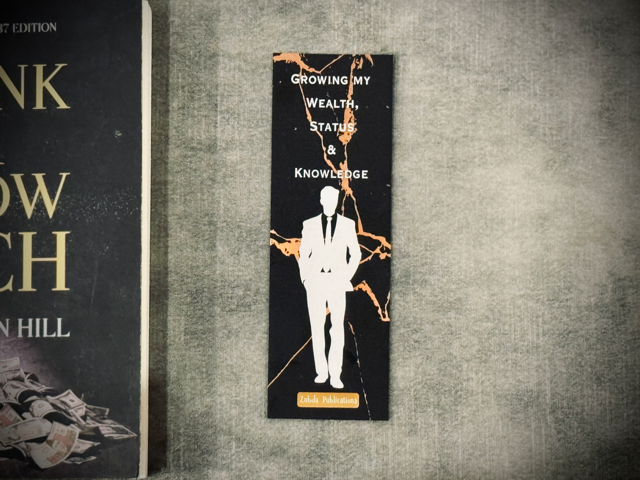 A laminated card-stock bookmark with black and gold marble with words 'growing my wealth, status and knowledge' placed on a surface with think and grow rich book in the background