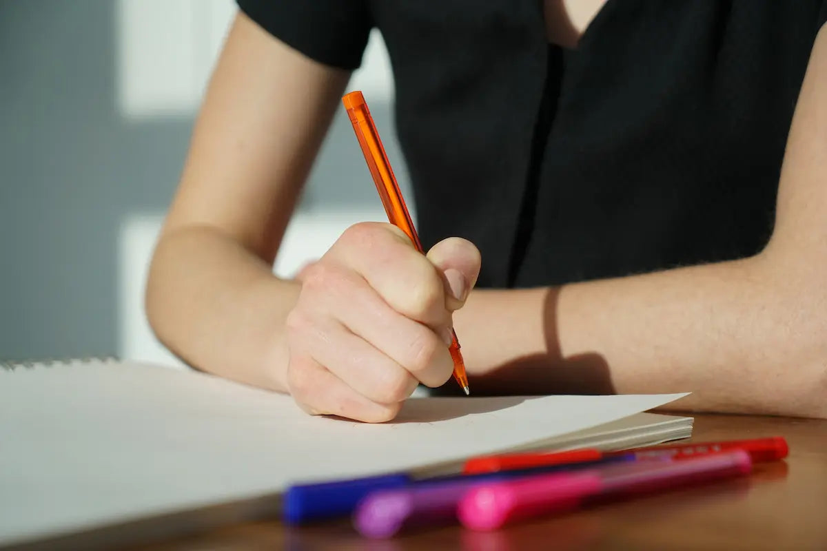 Person writing with an orange pen on a notebook, surrounded by colorful pens on a wooden surface.