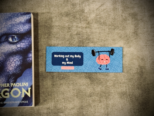 A laminated card stock bookmark with a cartoon brain lifting weight and a quote 'working out my body & my mind". Placed next to the book Eragon.
