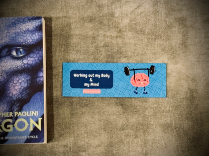 A laminated card stock bookmark with a cartoon brain lifting weight and a quote 'working out my body & my mind". Placed next to the book Eragon.